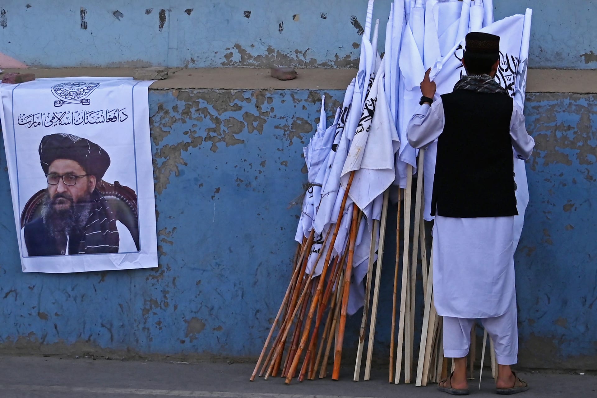   بائع أفغاني يفحص أعلام طالبان بجانب ملصق لزعيم طالبان عبد الغني بارادار على طول شارع في كابول - 27 أغسطس 2021 