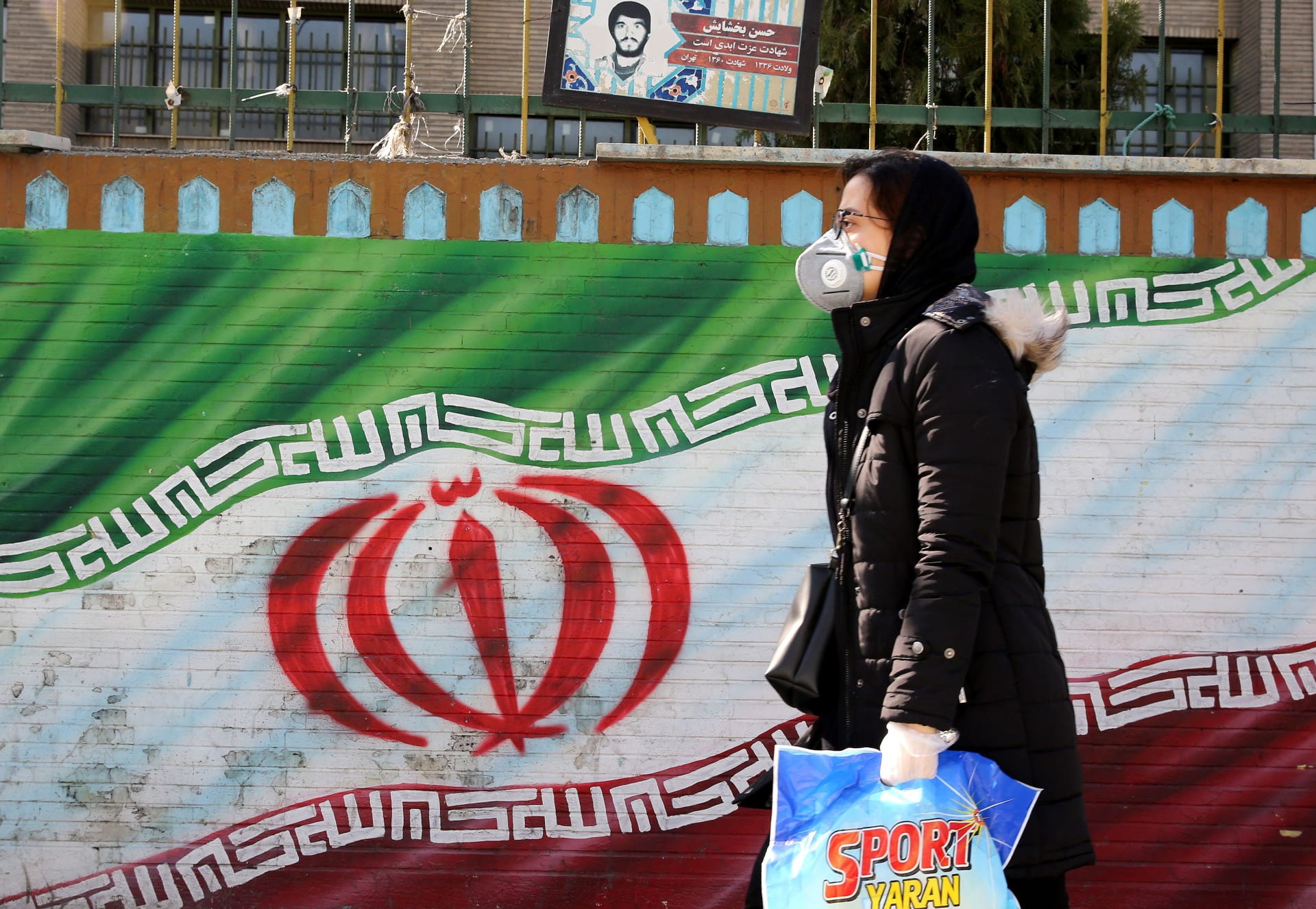 امرأة إيرانية ترتدي قناعًا تسير أمام لوحة جدارية تعرض علم بلادها في طهران، 4 مارس 2020